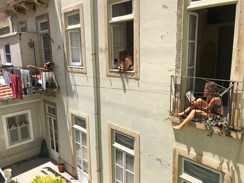 Zwei junge Frauen und  ein junger Mann sitzen in Badebekleidung  in den Fenstern ihrer Wohnungen und genießen die Sonne während des coronabedingten Lockdowns in Lissabon.